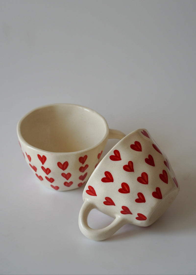 Two cream-colored ceramic mugs decorated with a pattern of small red hearts, displayed on a light gray background.