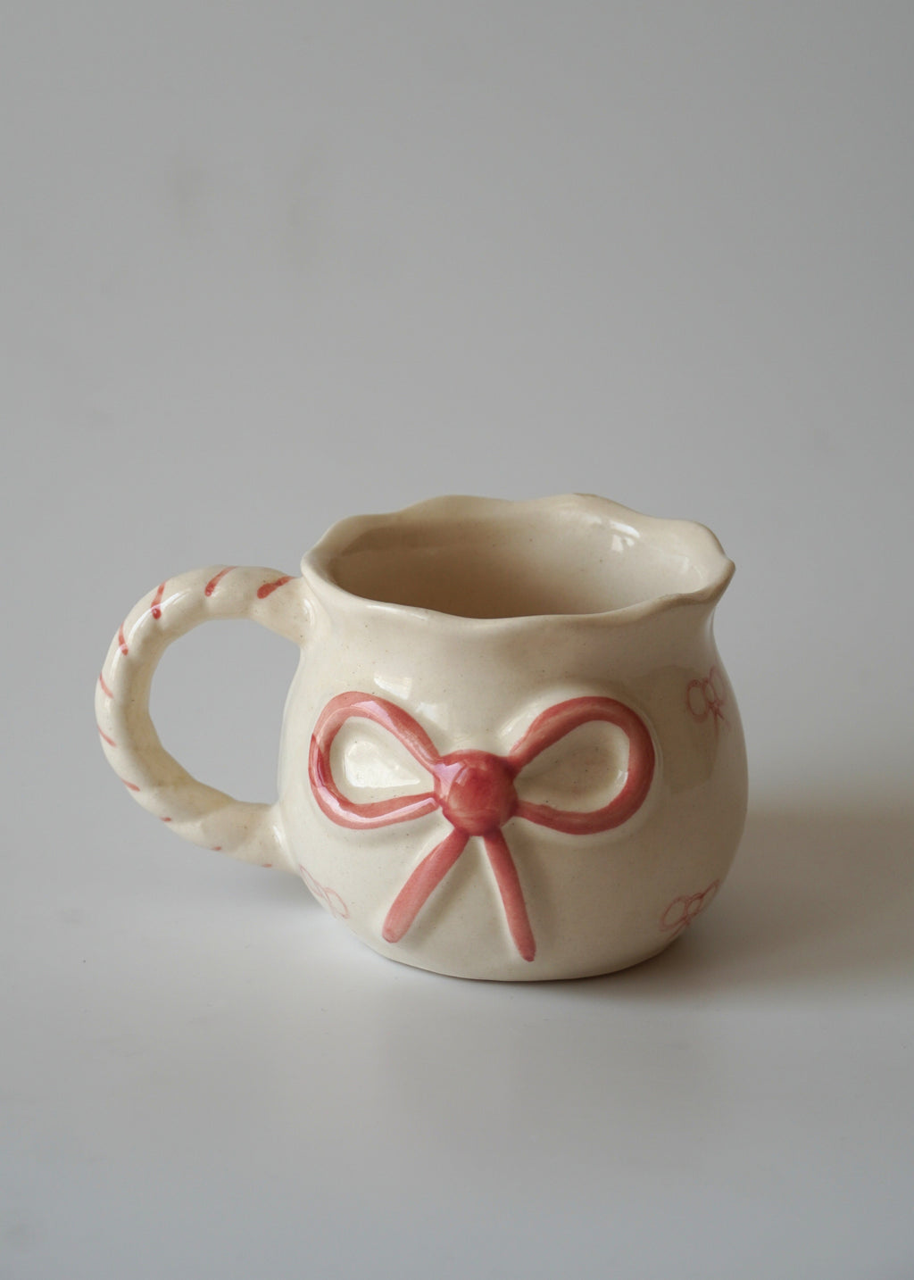 A small cream-colored ceramic mug with a pink bow design on the front and pink dashed lines along the handle, set against a plain light gray background.