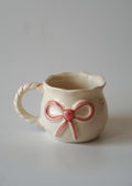 A small cream-colored ceramic mug with a pink bow design on the front and pink dashed lines along the handle, set against a plain light gray background.