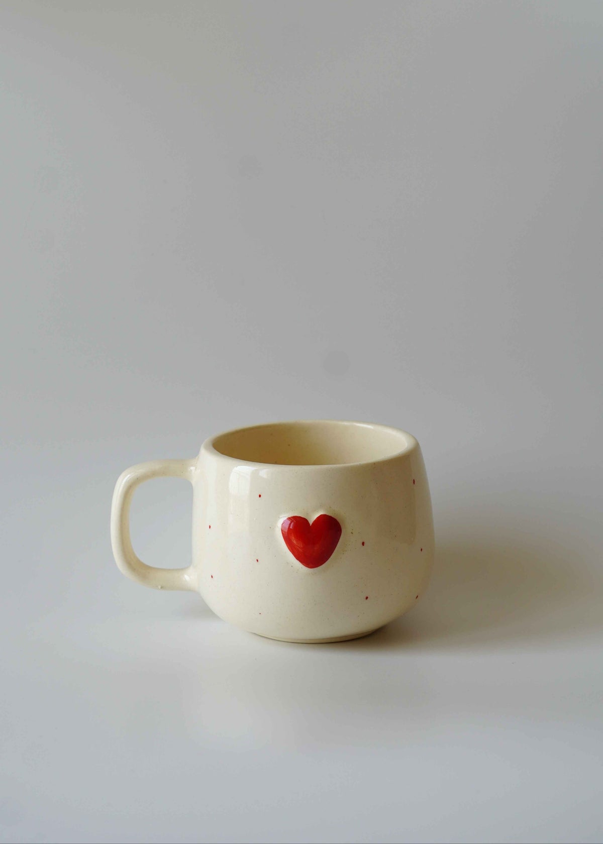 A cream-colored ceramic mug with a small red heart on the front, set against a plain light gray background.