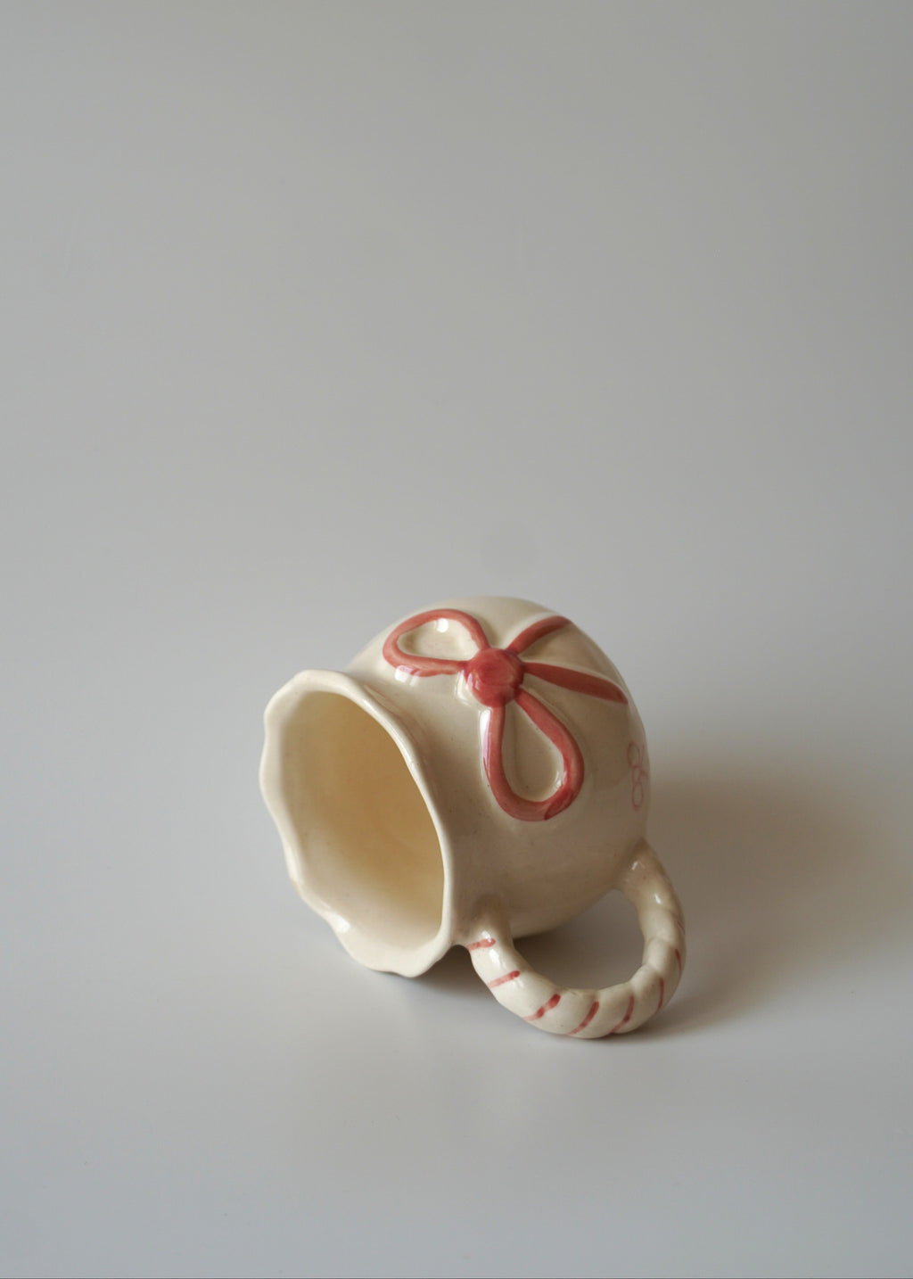 A small cream-colored ceramic mug with a handle shaped like a candy cane, decorated with a red bow and striped accents, photographed on a neutral light gray background.