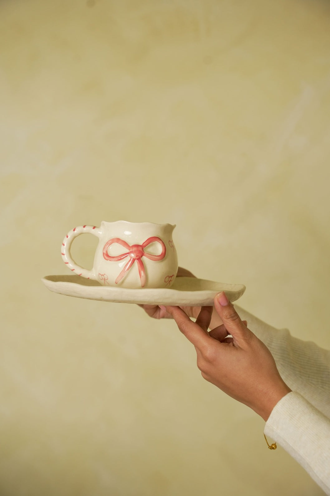 Hand holding a white teacup with a pink bow on a plate against a plain background