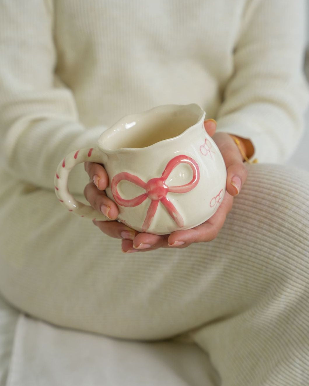 Person holding a ceramic mug with a pink bow design, wearing a beige sweater.