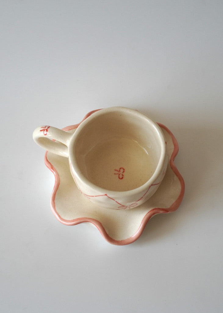 A small cream-colored ceramic teacup with a matching scalloped saucer, both decorated with delicate pink and red hand-painted details, set against a plain light gray background.