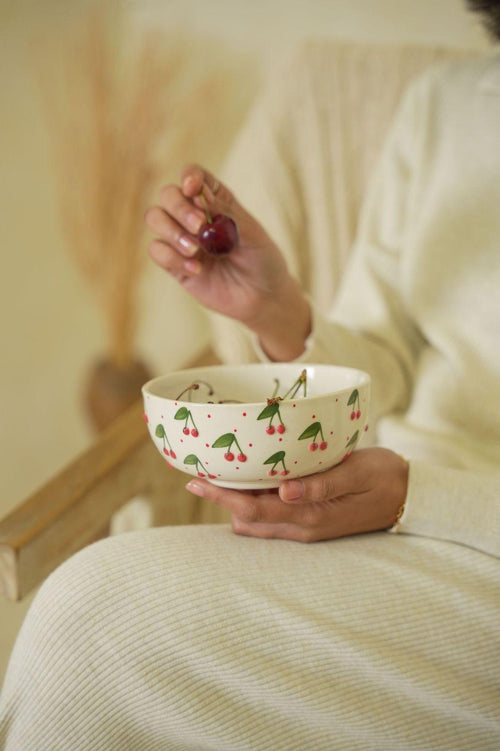 Person holding a bowl with cherry design and a cherry in their hand, sitting on a couch.