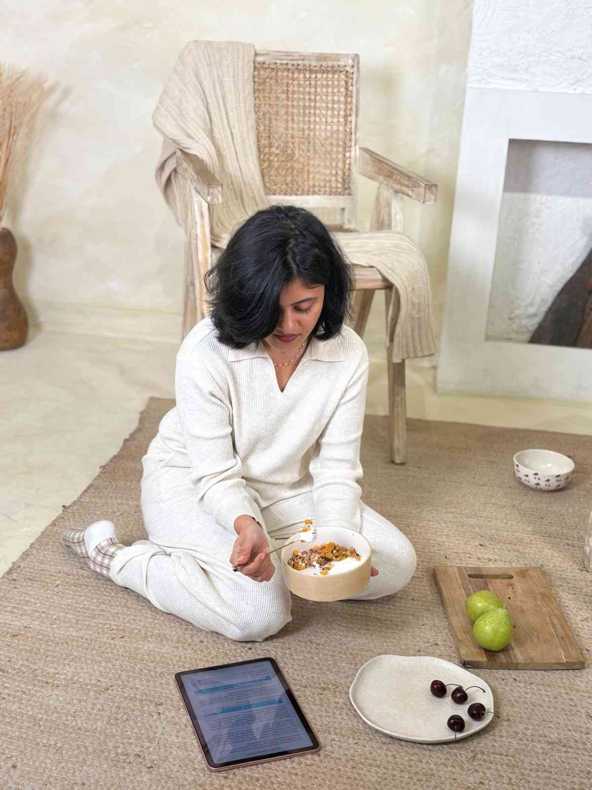 Founder of Sumeko sitting on the floor with sumeko bowl of food, tablet, and fruit in a cozy room.