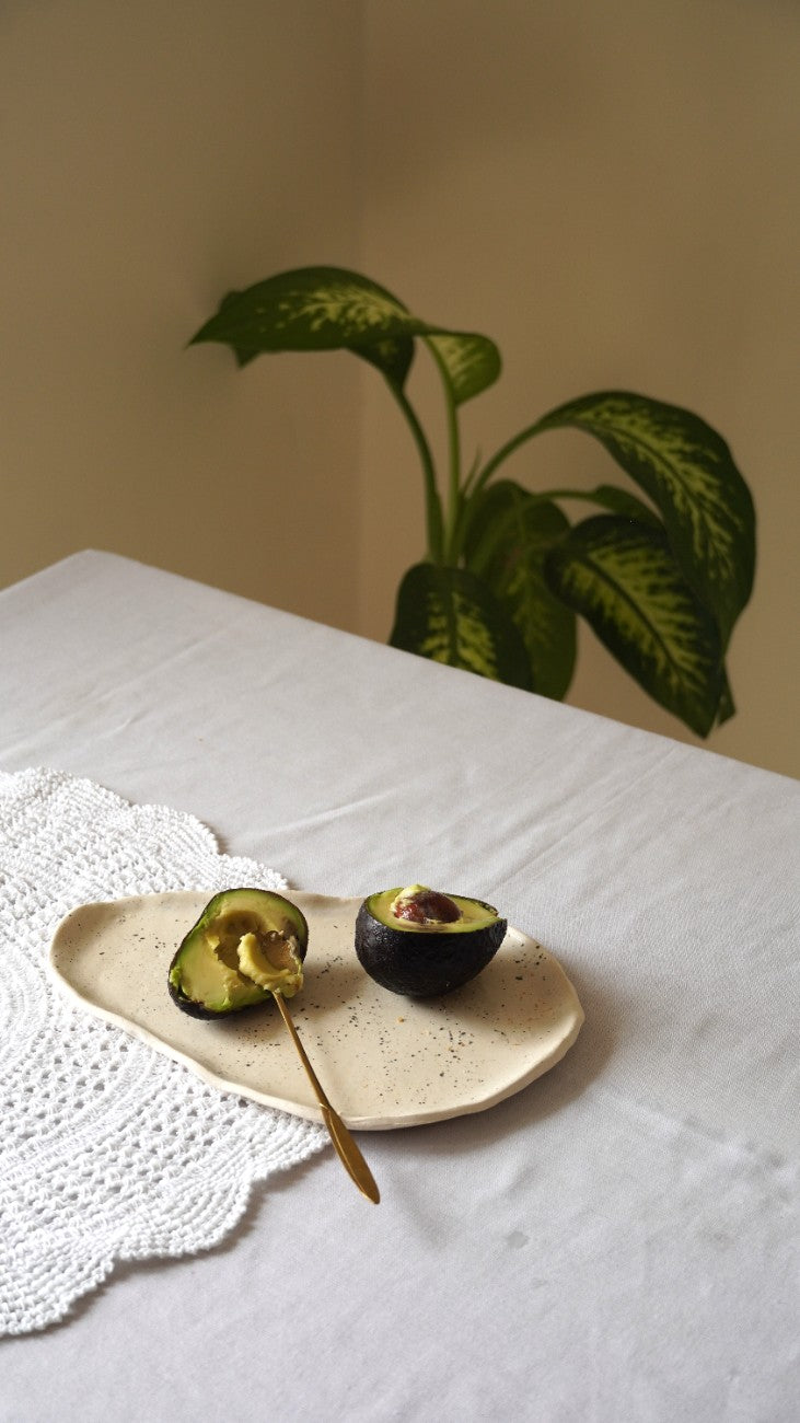 A ceramic speckled beige plate holding a halved avocado and a spoon, set on a white tablecloth next to a white lace doily, with a green houseplant in the background.