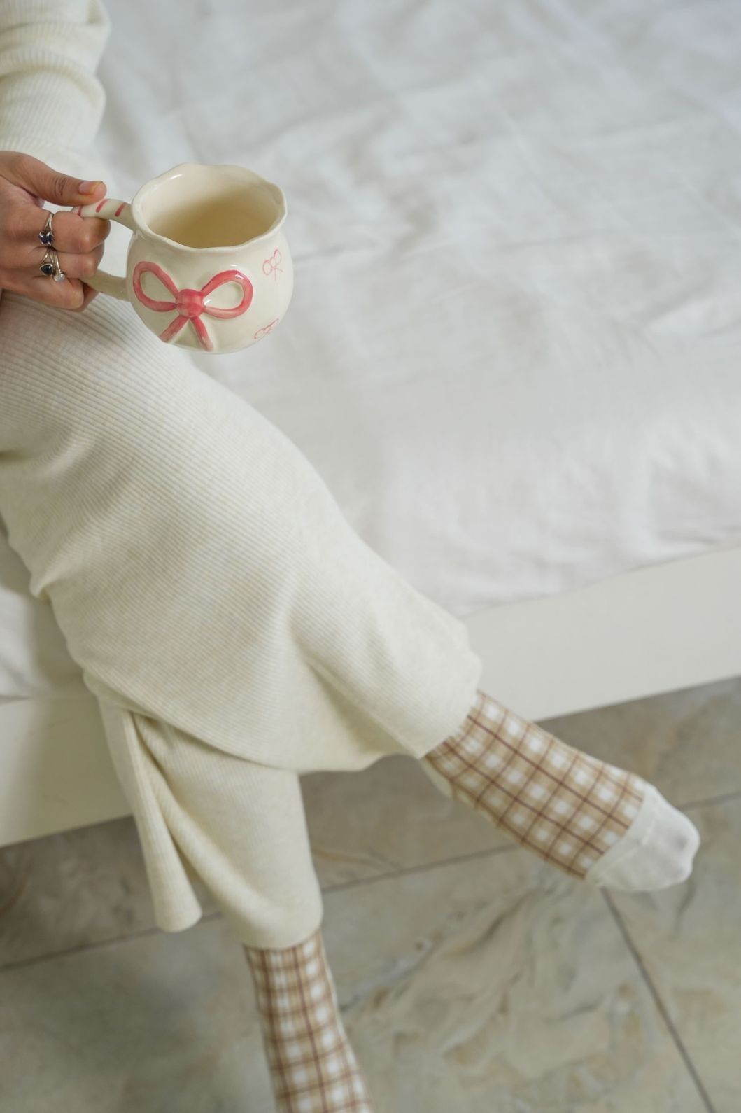 Person holding a mug with a bow design, wearing a white sweater and checkered socks.