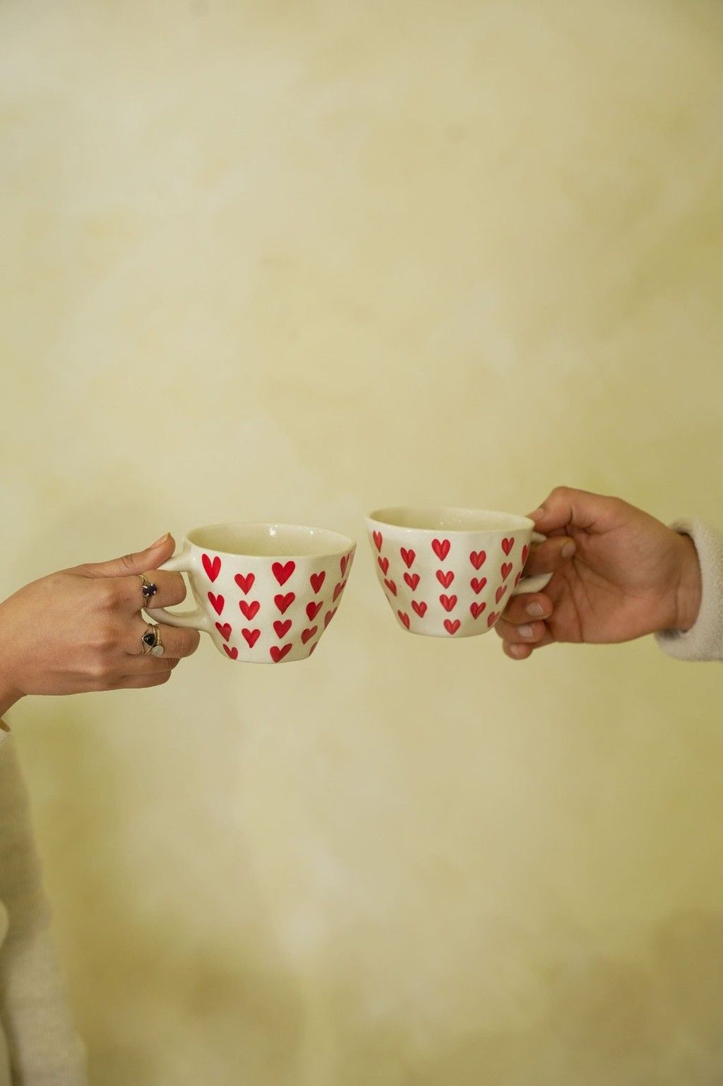 Two hands holding mugs with red heart patterns against a plain background