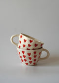 Two stacked ceramic mugs with a cream-colored glaze, decorated with a repeating pattern of small red hearts, set against a plain light gray background.