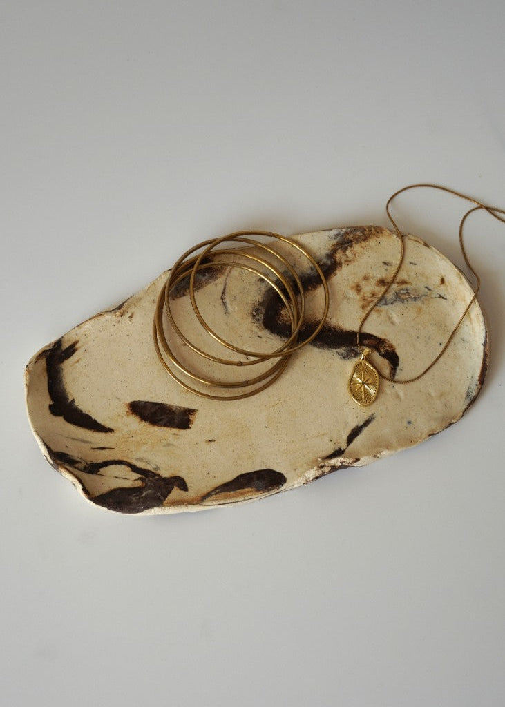 A set of gold jewelry, including stacked bangles and a necklace with a small round pendant, displayed on a rustic, irregularly shaped beige and brown ceramic tray against a plain light gray background.