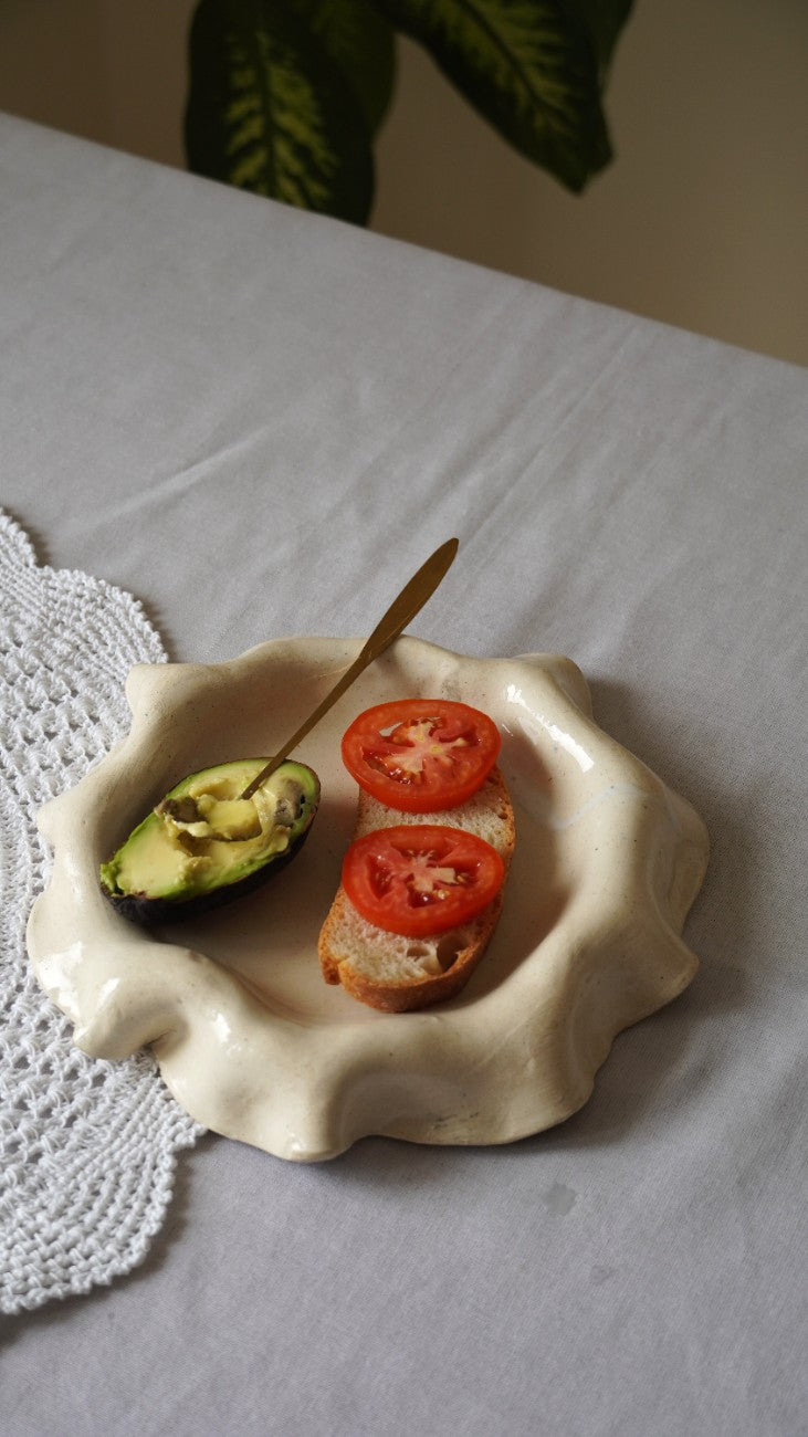 A scalloped cream-colored ceramic dish holding avocado halves and tomato slices on a white tablecloth, with a small spoon resting inside the dish.