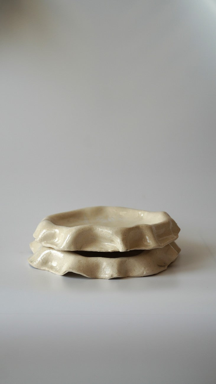 Two stacked, organic-shaped ceramic dishes in a warm cream or beige glaze, set against a plain light gray background.