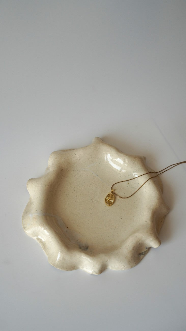 A small gold pendant necklace with a thin chain rests inside a shallow, organic-shaped cream ceramic dish on a plain light gray background.