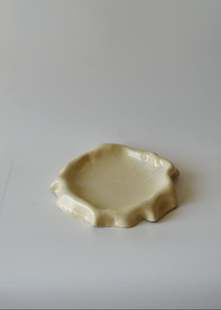 A small, organic-shaped ceramic dish in a light cream or pale yellow glaze, set against a plain light gray background.