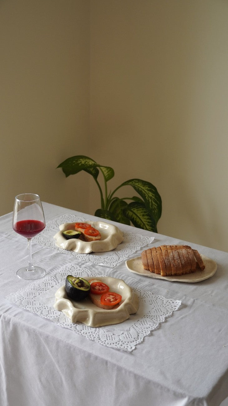 Three round ceramic plates with organic, shell-like edges are arranged on a white tablecloth. One plate holds sliced avocado and tomato, another holds a slice of bread, and a third is empty.