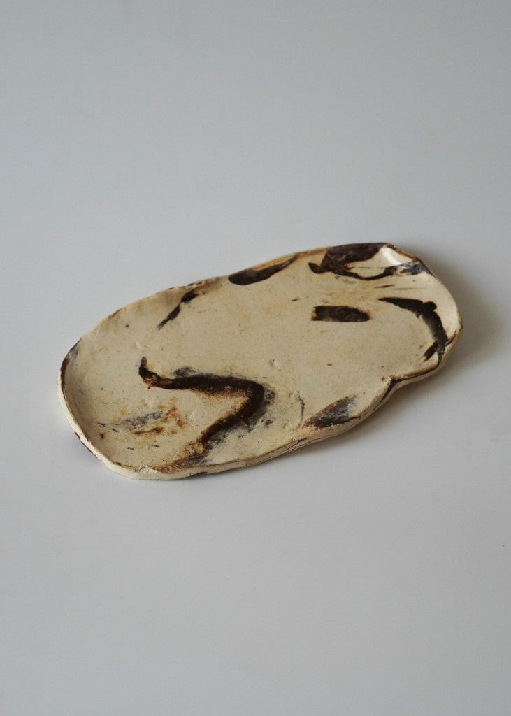 An organic-shaped ceramic plate with a cream-colored glaze and dark brown abstract painted patterns, photographed on a neutral light gray background.