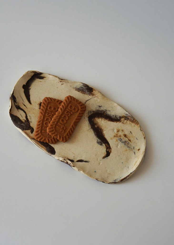 A small irregular ceramic plate with a cream and dark brown marbled pattern, holding two brown sugar cookies, photographed on a light gray background.
