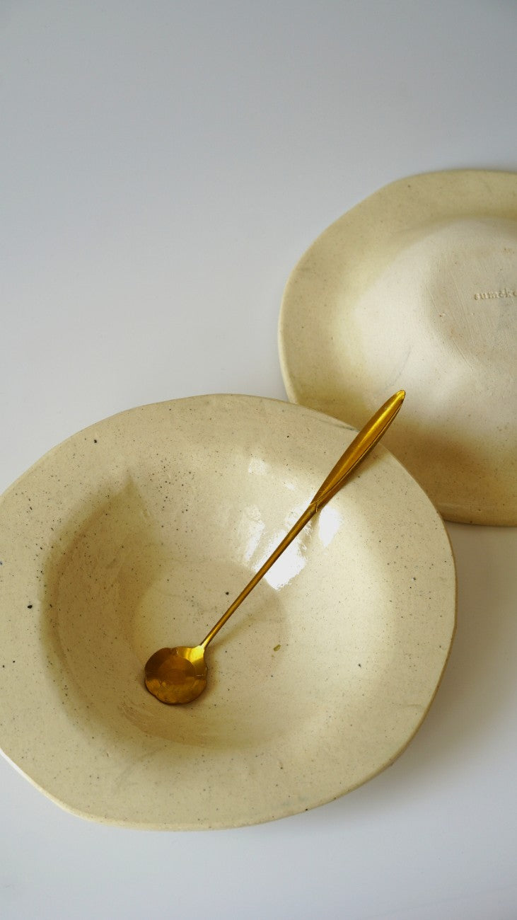 A beige speckled ceramic bowl with a gold spoon inside, with another matching bowl partially visible in the background on a light gray surface.