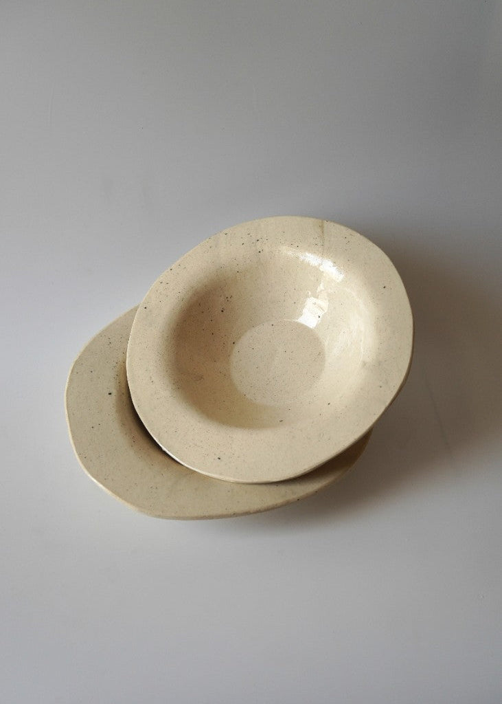Two small, handcrafted ceramic bowls in a speckled cream-beige glaze, stacked on a neutral light gray background.