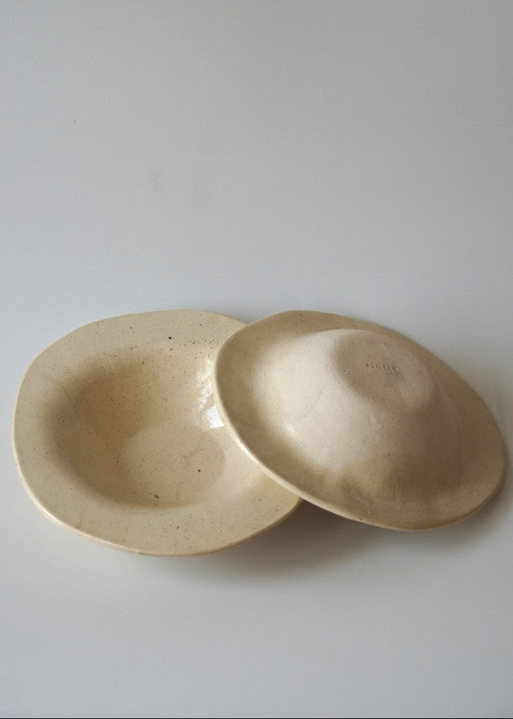 Two small shallow ceramic dishes or bowls in a warm off-white/cream glaze, one standing and one lying on its side, photographed on a plain light background.