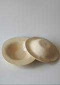 Two small shallow ceramic dishes or bowls in a warm off-white/cream glaze, one standing and one lying on its side, photographed on a plain light background.