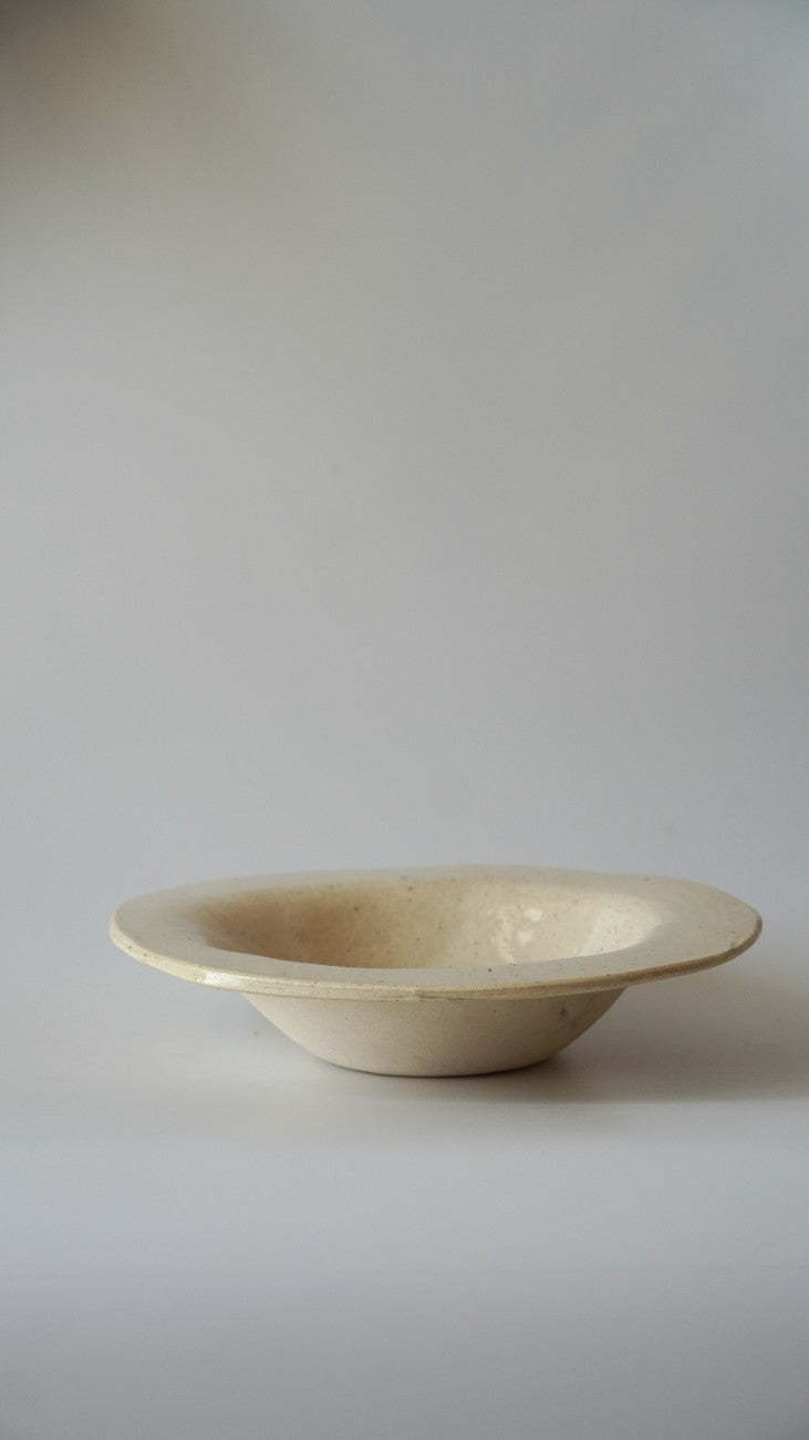 A small, shallow cream-colored ceramic bowl with a wide, slightly flared rim, photographed against a neutral light gray background.