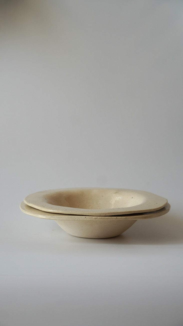 A pair of stacked, shallow ceramic bowls in a matte cream or off-white glaze, set against a plain light gray background.