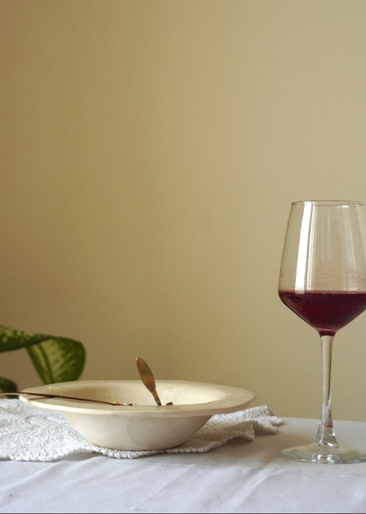 A beige ceramic bowl with a gold spoon resting inside, set on a white textured cloth next to a glass of red wine on a white tablecloth against a warm beige wall.