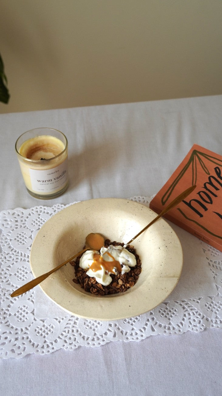 A styled tabletop scene with a cream-colored ceramic bowl containing a dessert topped with whipped cream and caramel sauce, alongside a gold spoon. A lit candle in a glass jar and an orange book or notebook are placed nearby on a white lace doily over a white tablecloth.