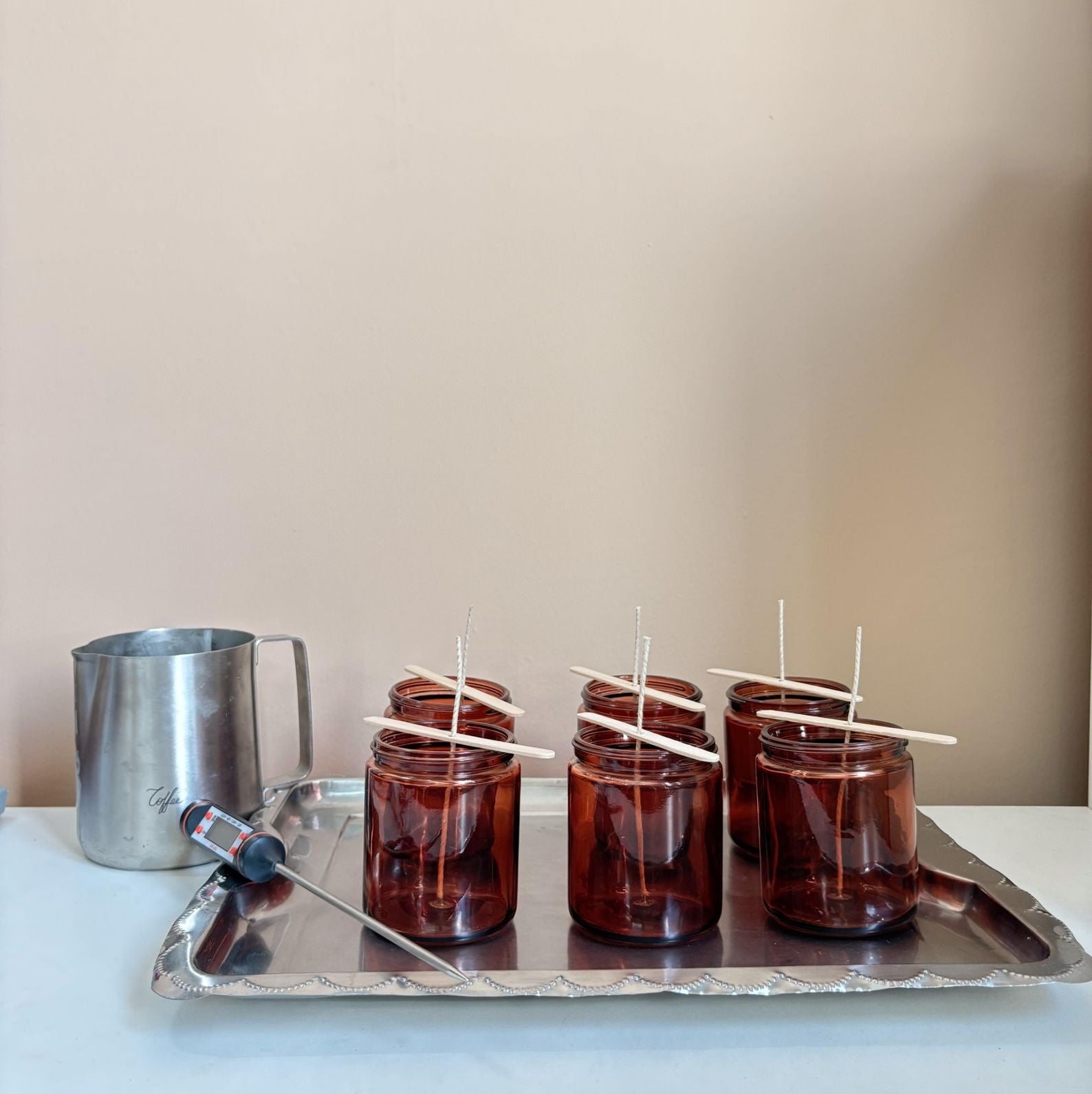 Brown glass jars with sticks on a metal tray next to a silver pitcher against a beige wall.