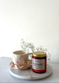 Candle labeled 'warm vanilla' next to a mug on a white surface with a white background