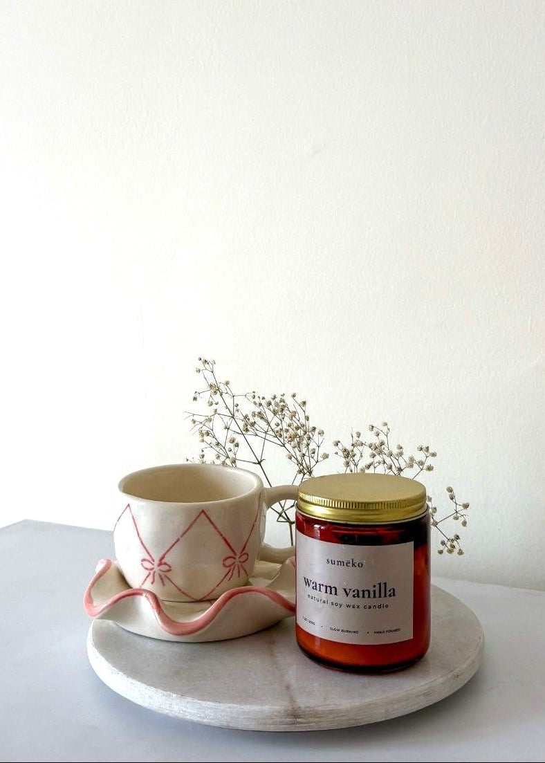 Candle labeled 'warm vanilla' next to a mug on a white surface with a white background