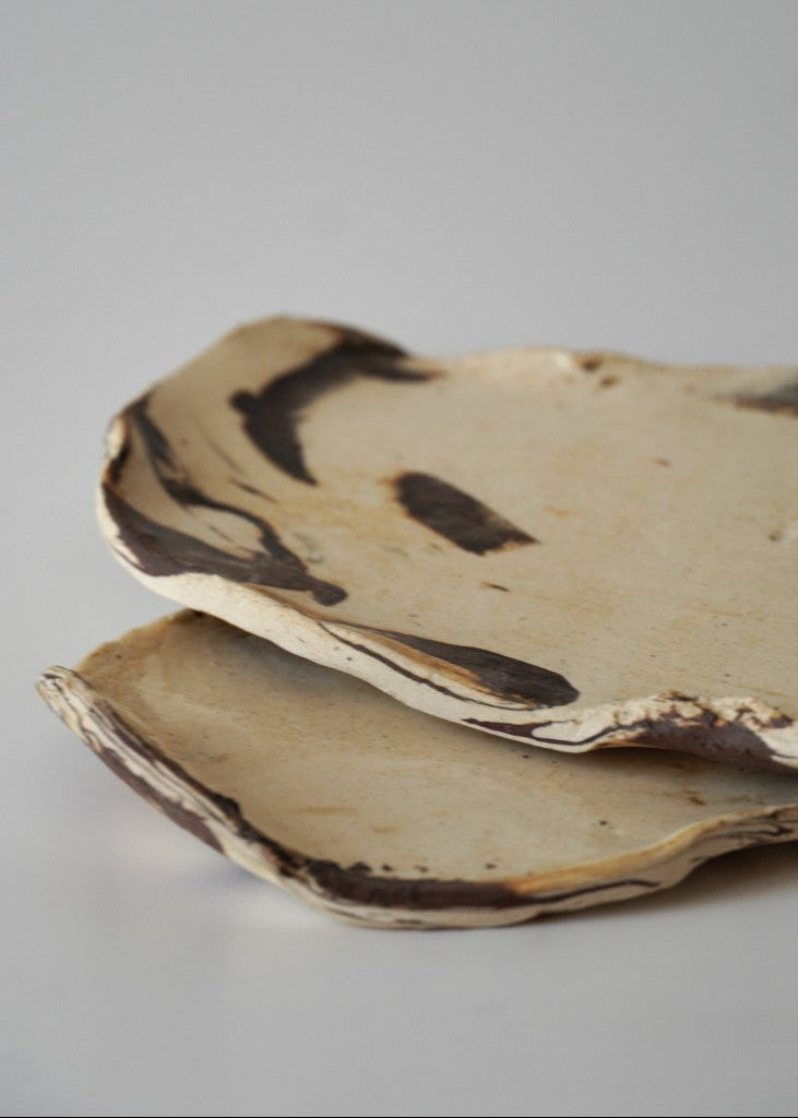 Two handmade ceramic plates with organic, irregular shapes and dark brown painted accents, stacked on a light gray background.