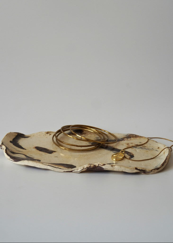 A set of gold jewelry, including bangles and a necklace with a small heart pendant, displayed on a rustic, irregularly shaped ceramic plate against a plain light gray background.