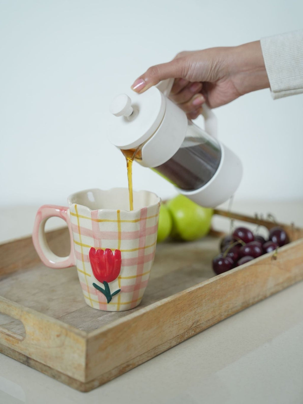 Person pouring coffee from a white French press into a mug with a floral design on a wooden tray.