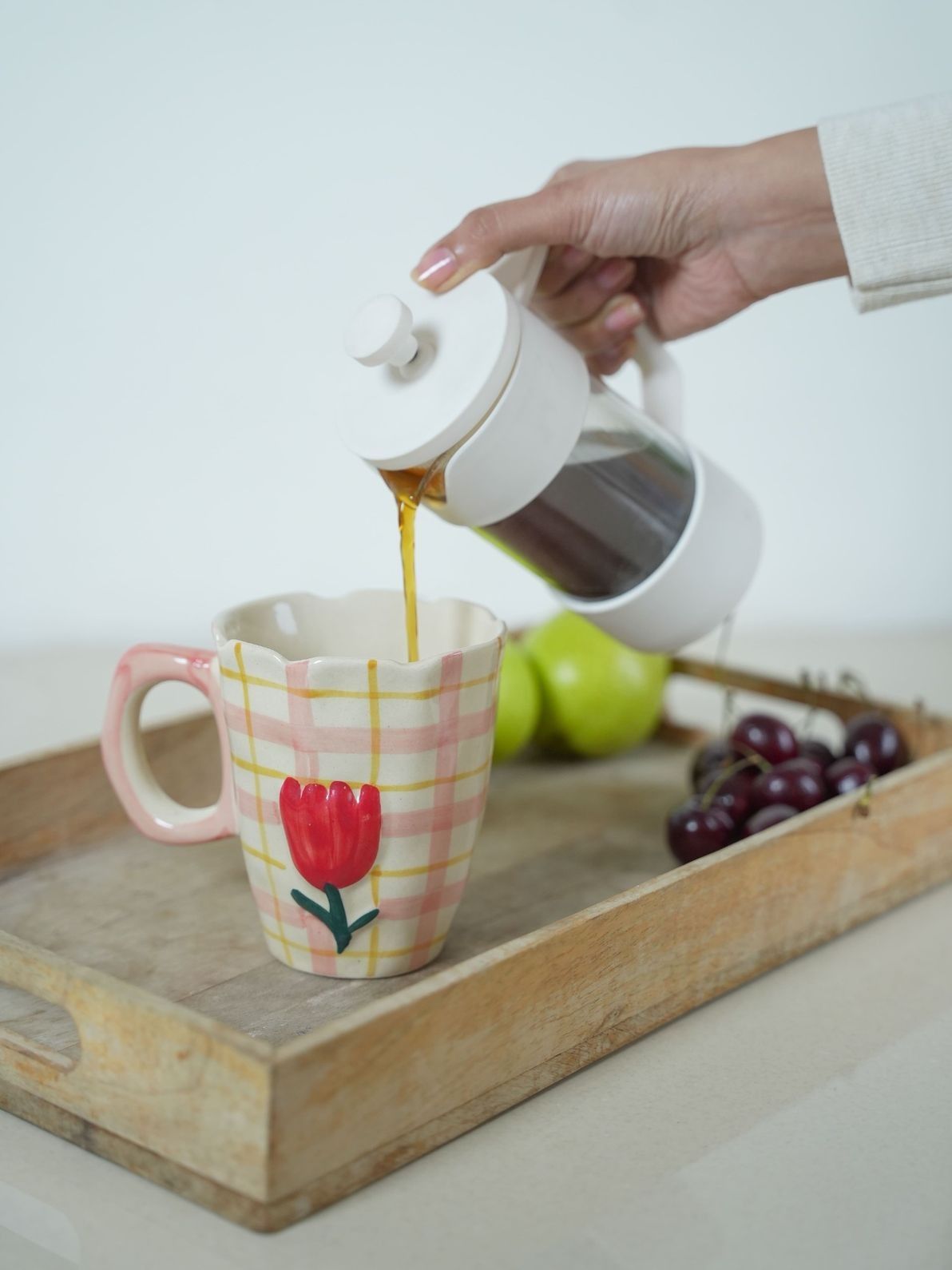Person pouring coffee from a white French press into a mug with a floral design on a wooden tray.