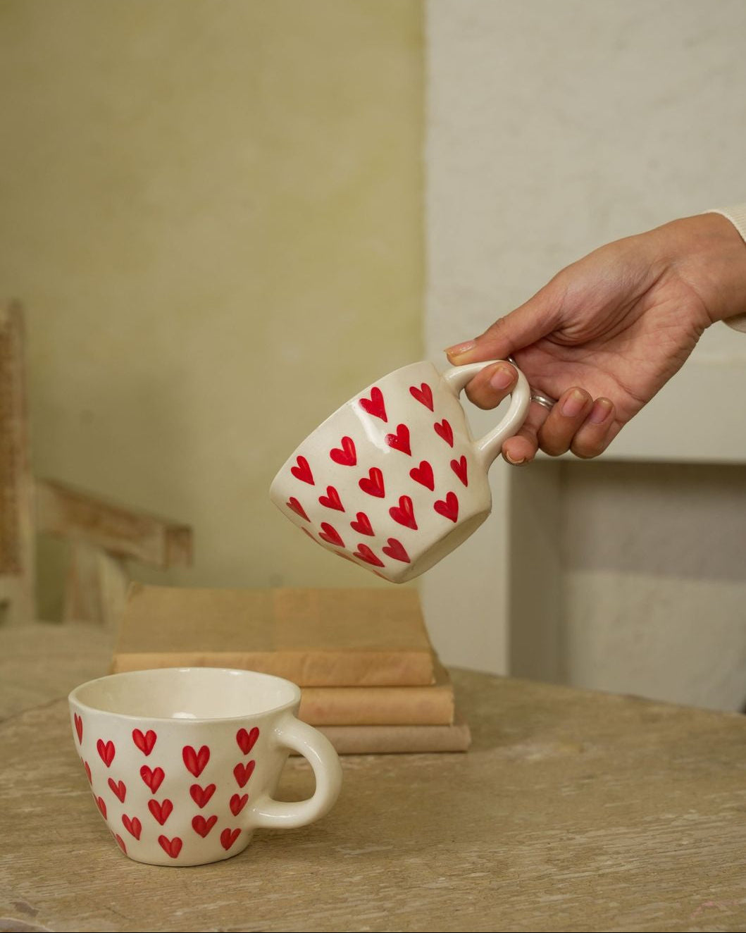 Hand holding a ceramic mug with red heart patterns over another similar mug on a wooden surface.